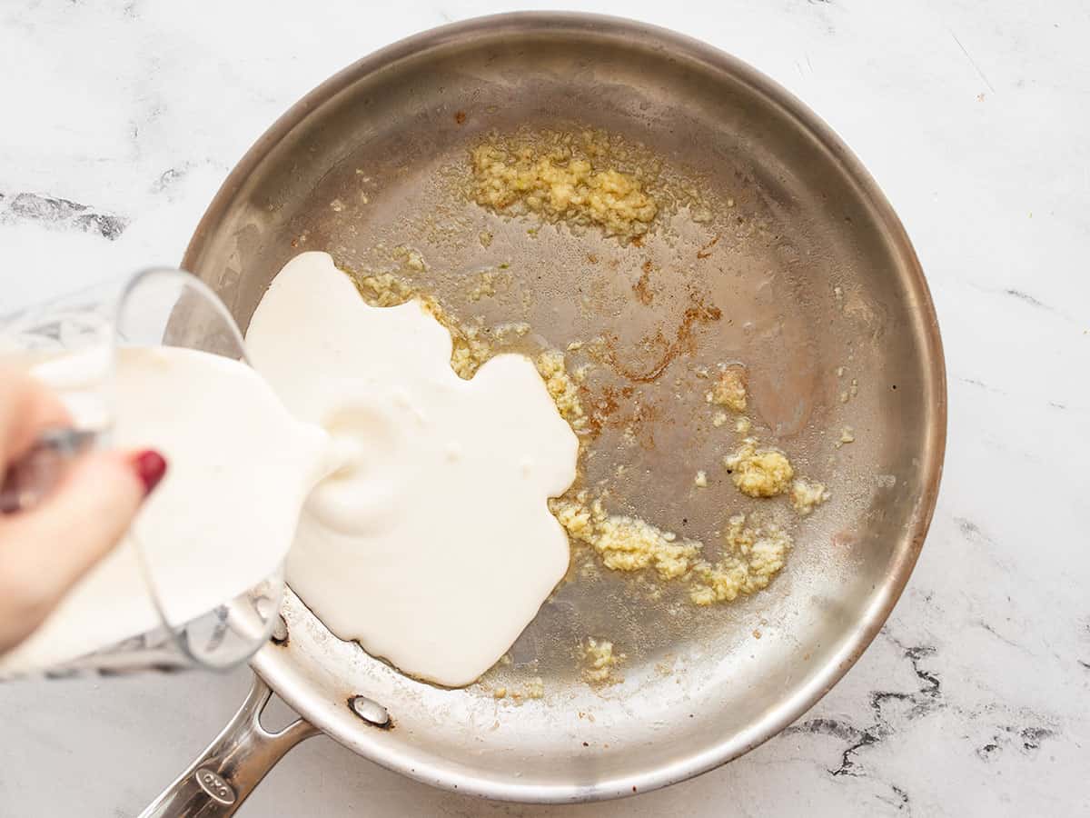 Heavy cream being poured into a skillet with garlic