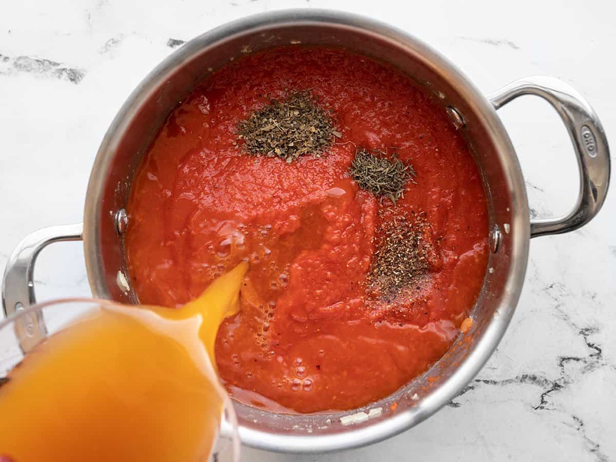 Peppers back in the soup pot with tomatoes, herbs and spices, and vegetable broth being poured into the pot.