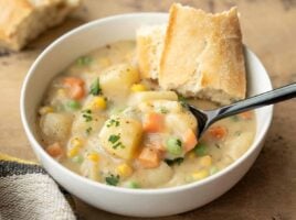 Side view of a bowl of vegetables with gravy with a piece of bread and a spoon.