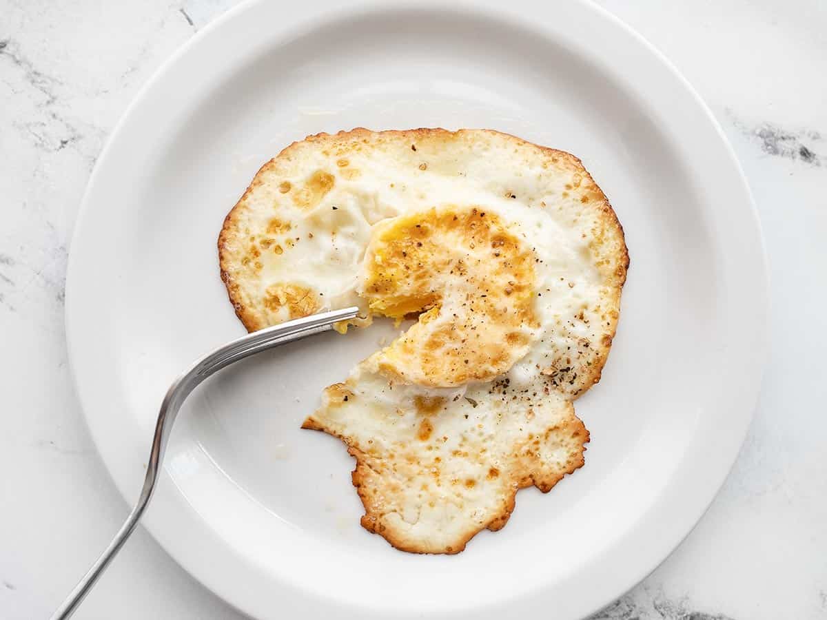 An egg cooked over hard on a plate with a fork piercing the cooked yolk.