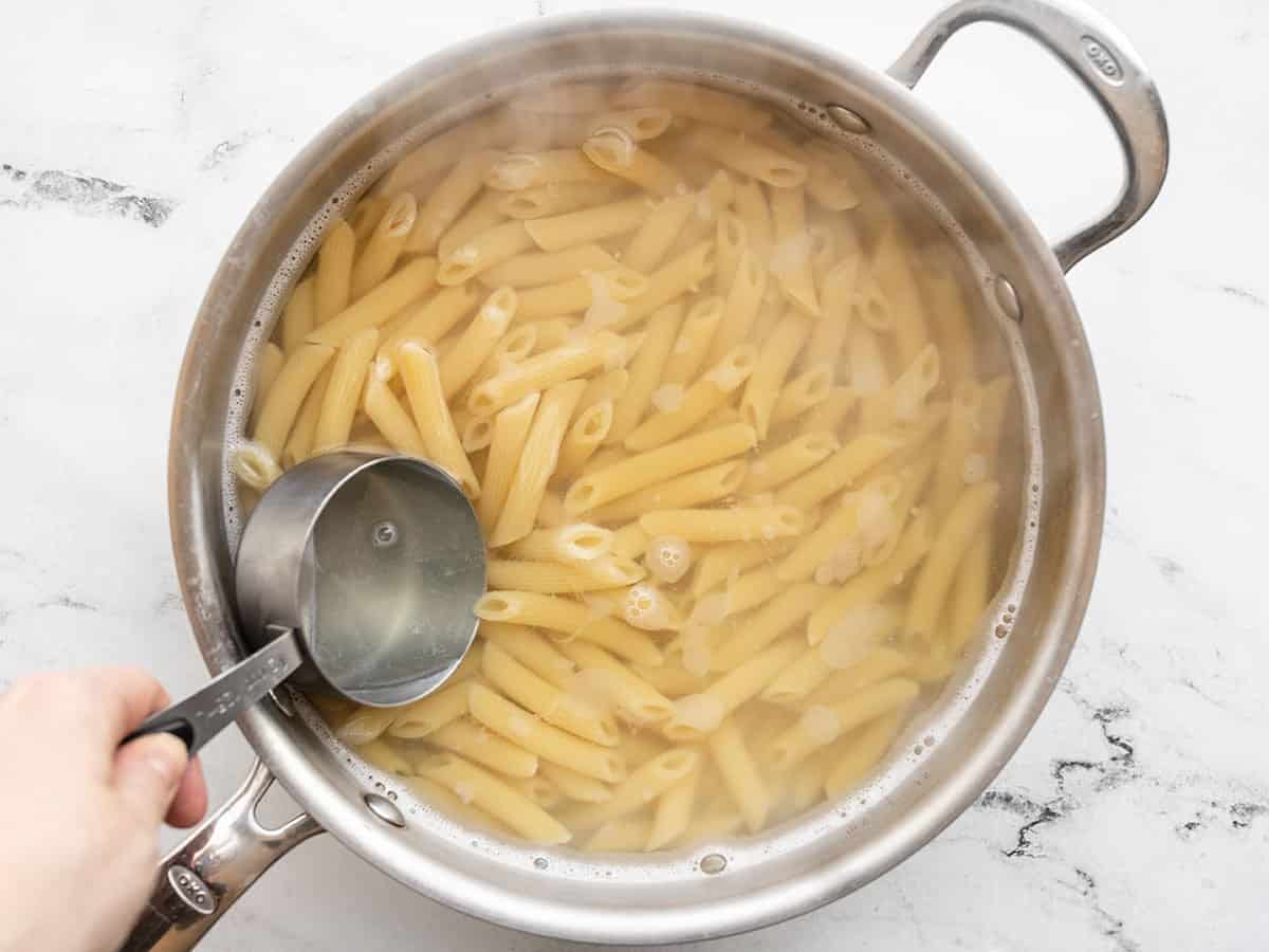 Boiled pasta in a pot, water being scooped out with a measuring cup.