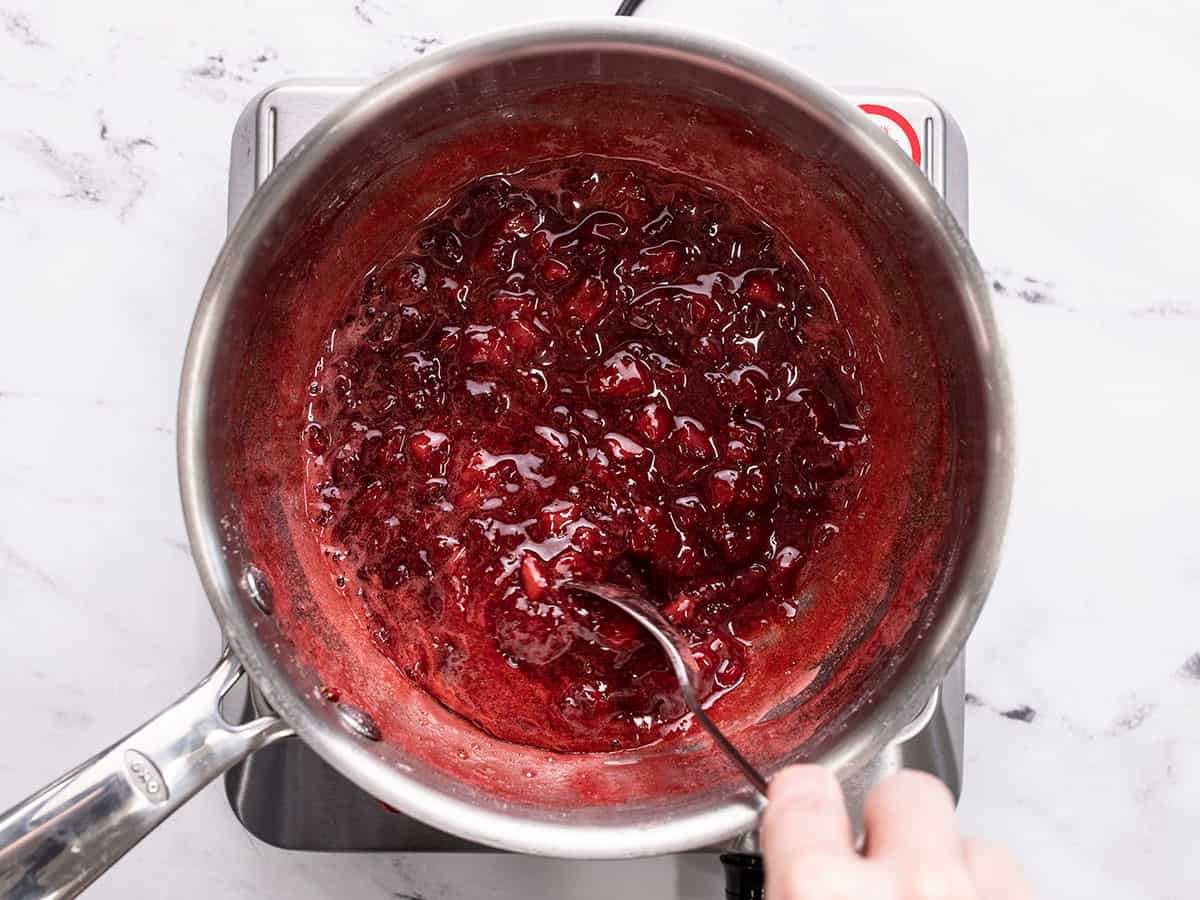 Thickened strawberries in the pot being stirred with a spoon.