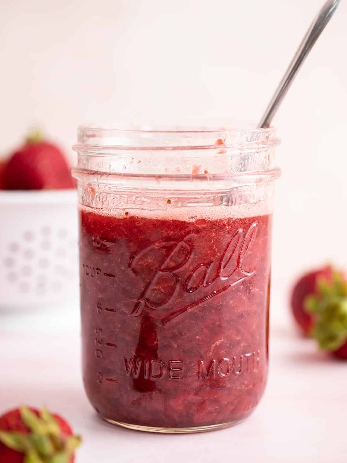 Side view of a jar of strawberry syrup with a spoon.