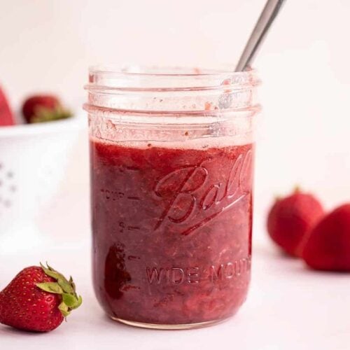 Side view of a jar of strawberry syrup with a spoon.