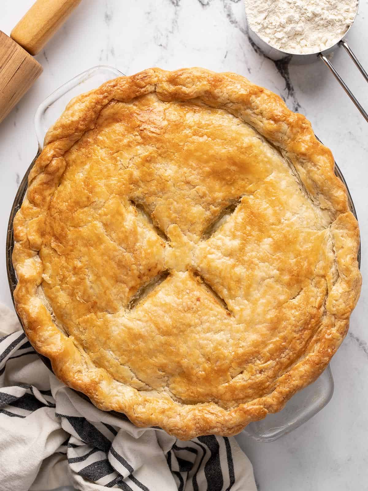 Overhead shot of a fully baked pie with a napkin, cup of flour, and rolling pin. surrounding it