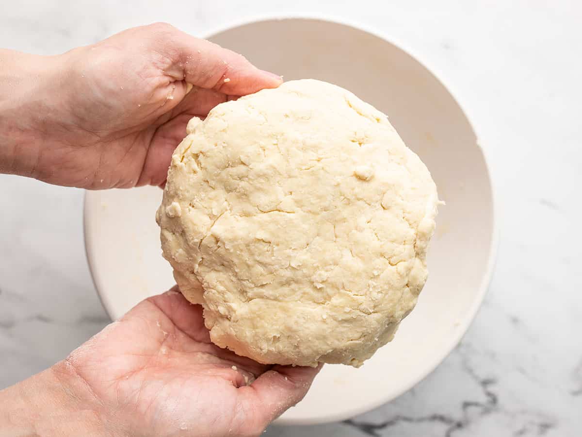 Overhead shot of two hands holding dough shaped in a flat disc over a white bowl.