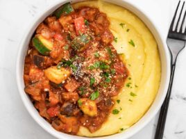 Close up overhead view of cornmeal and vegetables in red sauce in a bowl with a fork on the side.