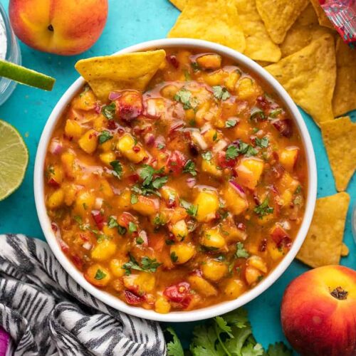 Overhead view of a bowl full of peach salsa surrounded by chips, peaches, and limes.