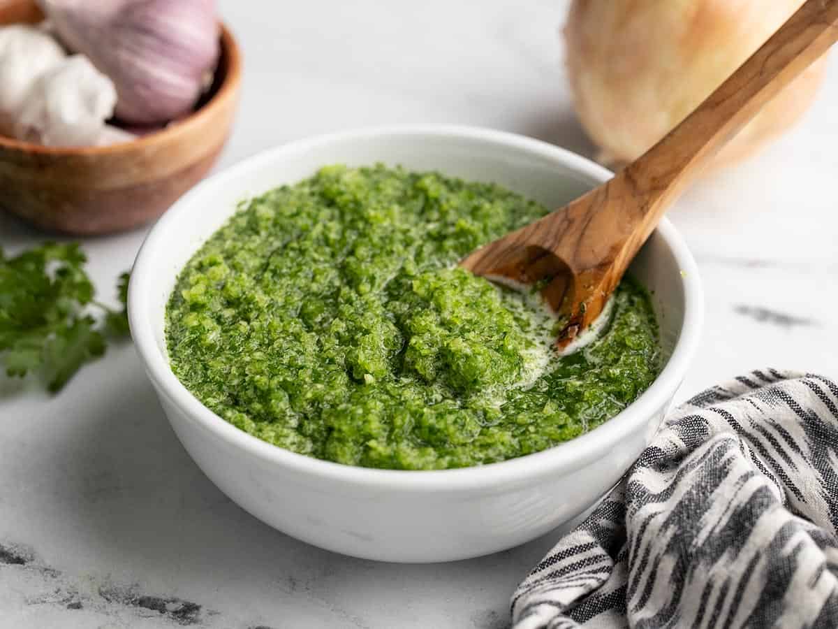 Side view of sofrito in a white bowl with a wood spoon in it and a black and white linen napkin next to it.