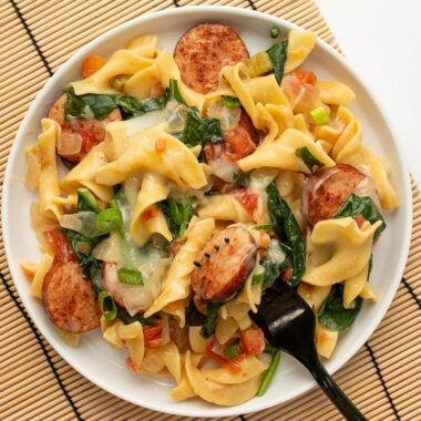 Cheesy sausage pasta on a plate with a black fork, on a bamboo mat.