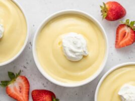 Overhead shot of vanilla pudding in a white bowl topped with a dollop of whipped cream and surrounded by strawberries.