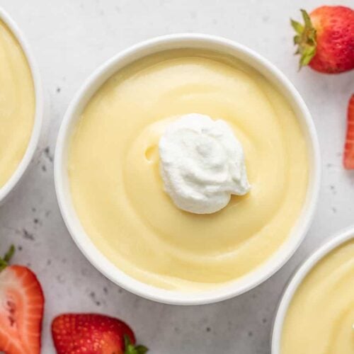 Overhead shot of vanilla pudding in a white bowl topped with a dollop of whipped cream and surrounded by strawberries.