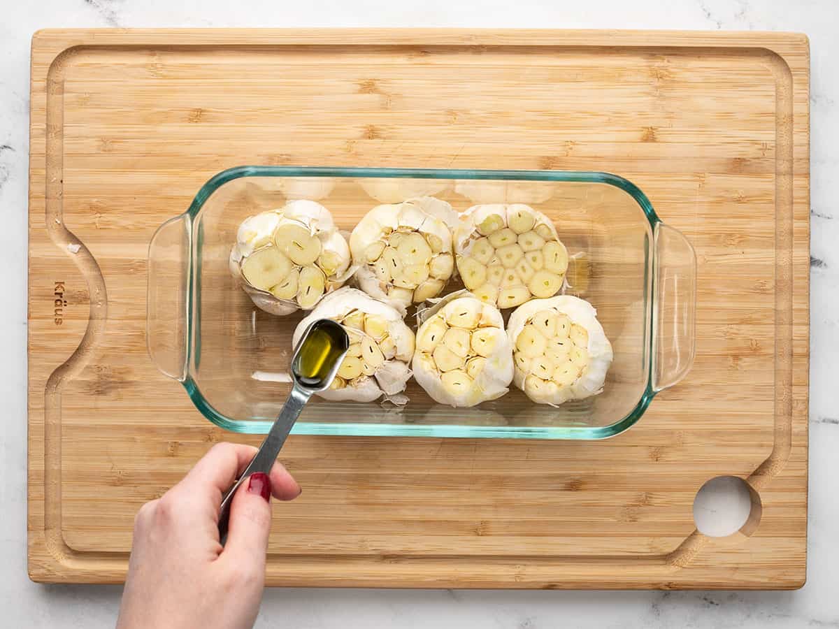 Oil being drizzled over garlic cloves in a glass dish.