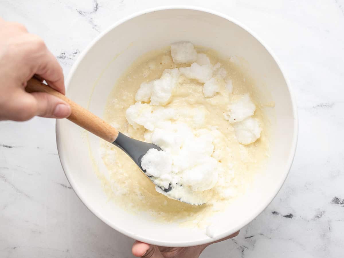 Overhead shot of egg whites being mixed into pancake batter in a white bowl with a wood handled rubber spatula in it.