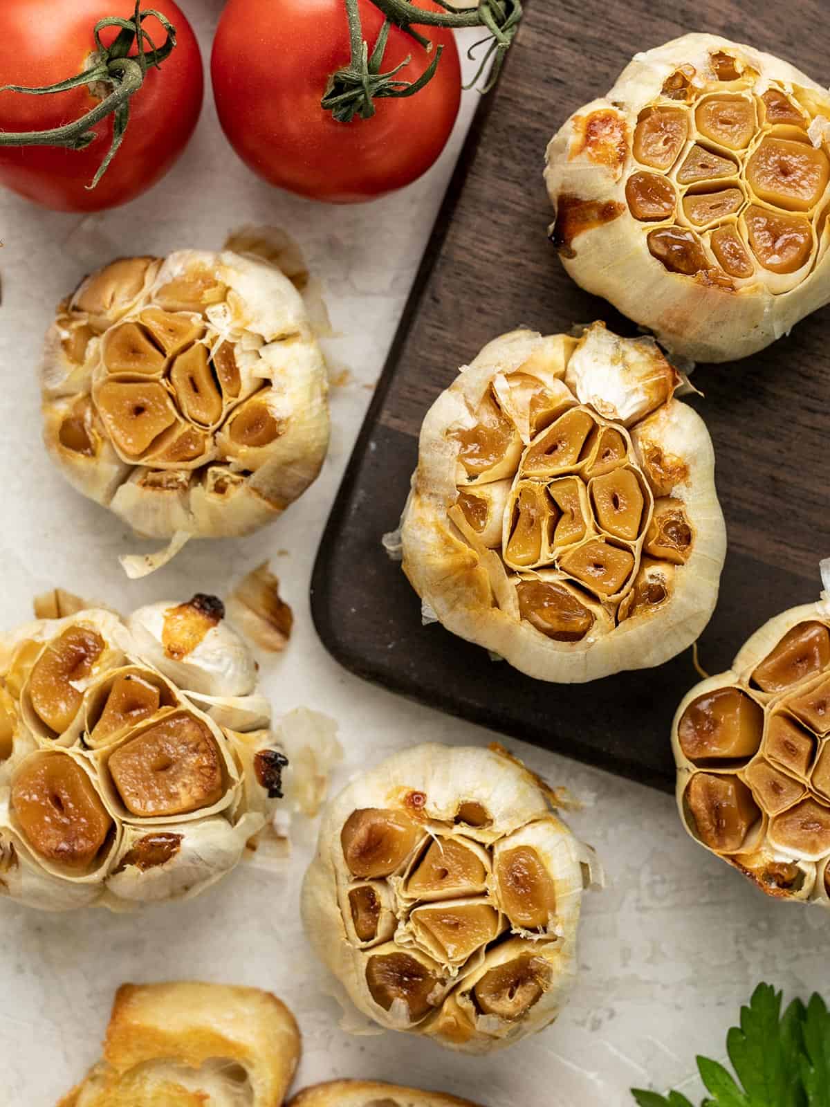 Roasted garlic heads scattered on a surface next to tomatoes.