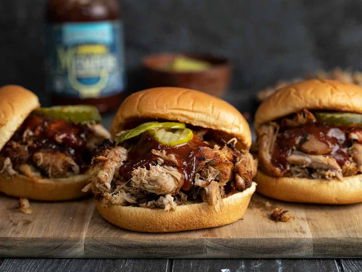 Three slow cooker pulled pork sandwiches on a wooden cutting board.