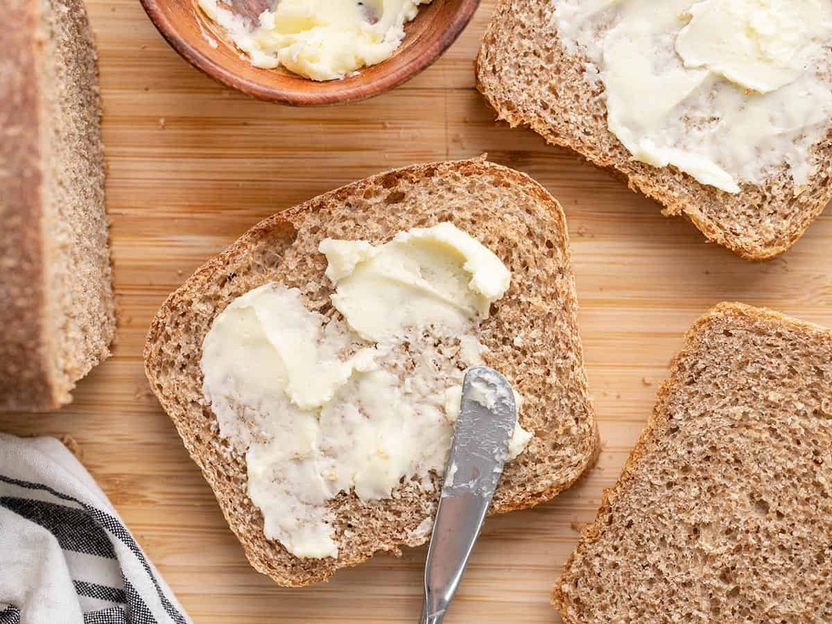 Overhead view of butter being spread on slices of wheat bread.