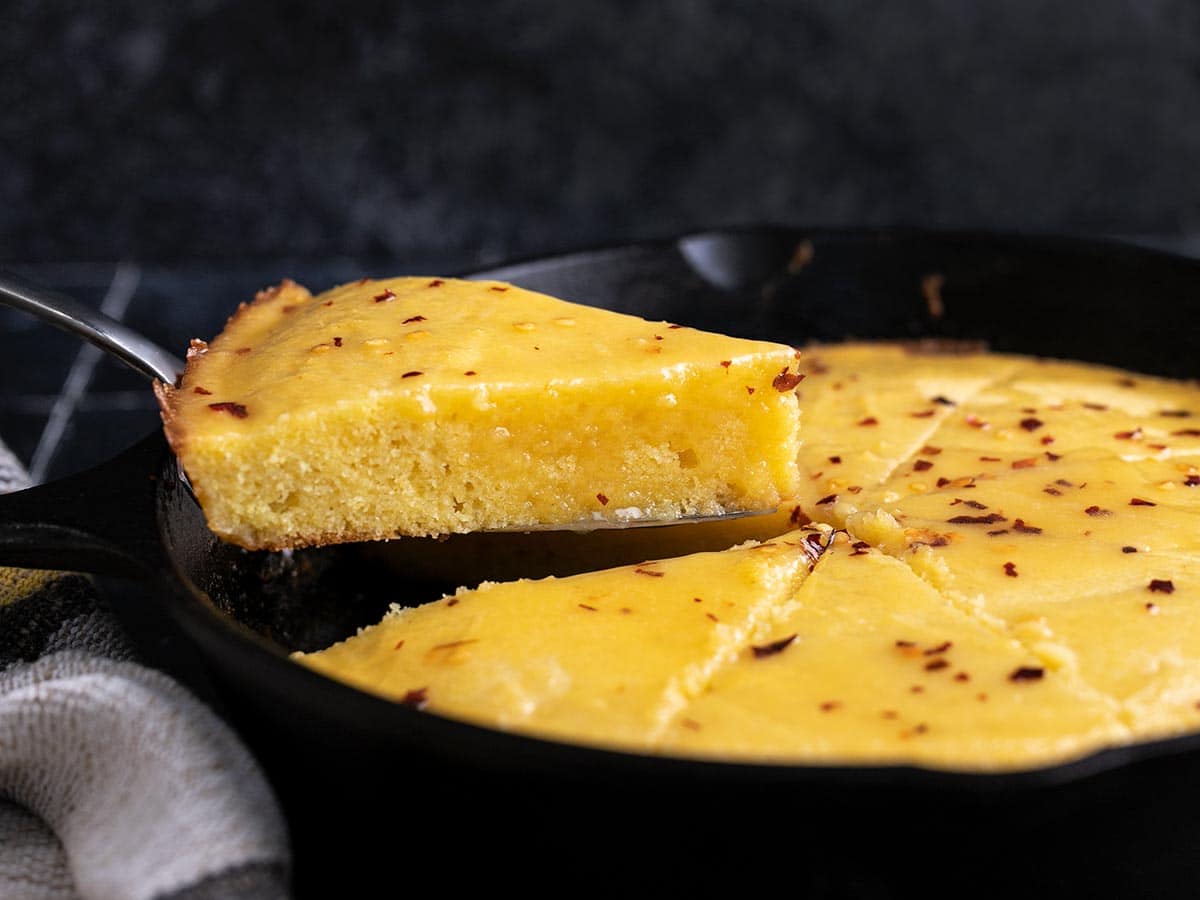 Side shot of hot honey cornbread in a cast iron skillet with a wedge being taken out.