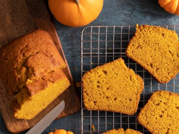 Overhead shot of sliced pumpkin bread with two mini pumpkins next to it on a dark background.