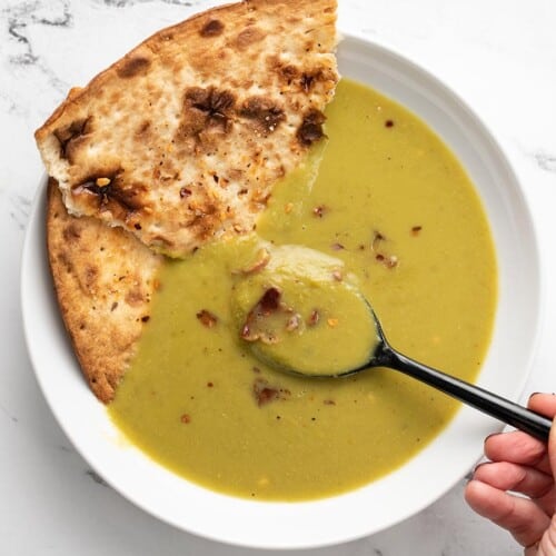 A spoon dipping into a bowl of pea soup with flatbread on the side.