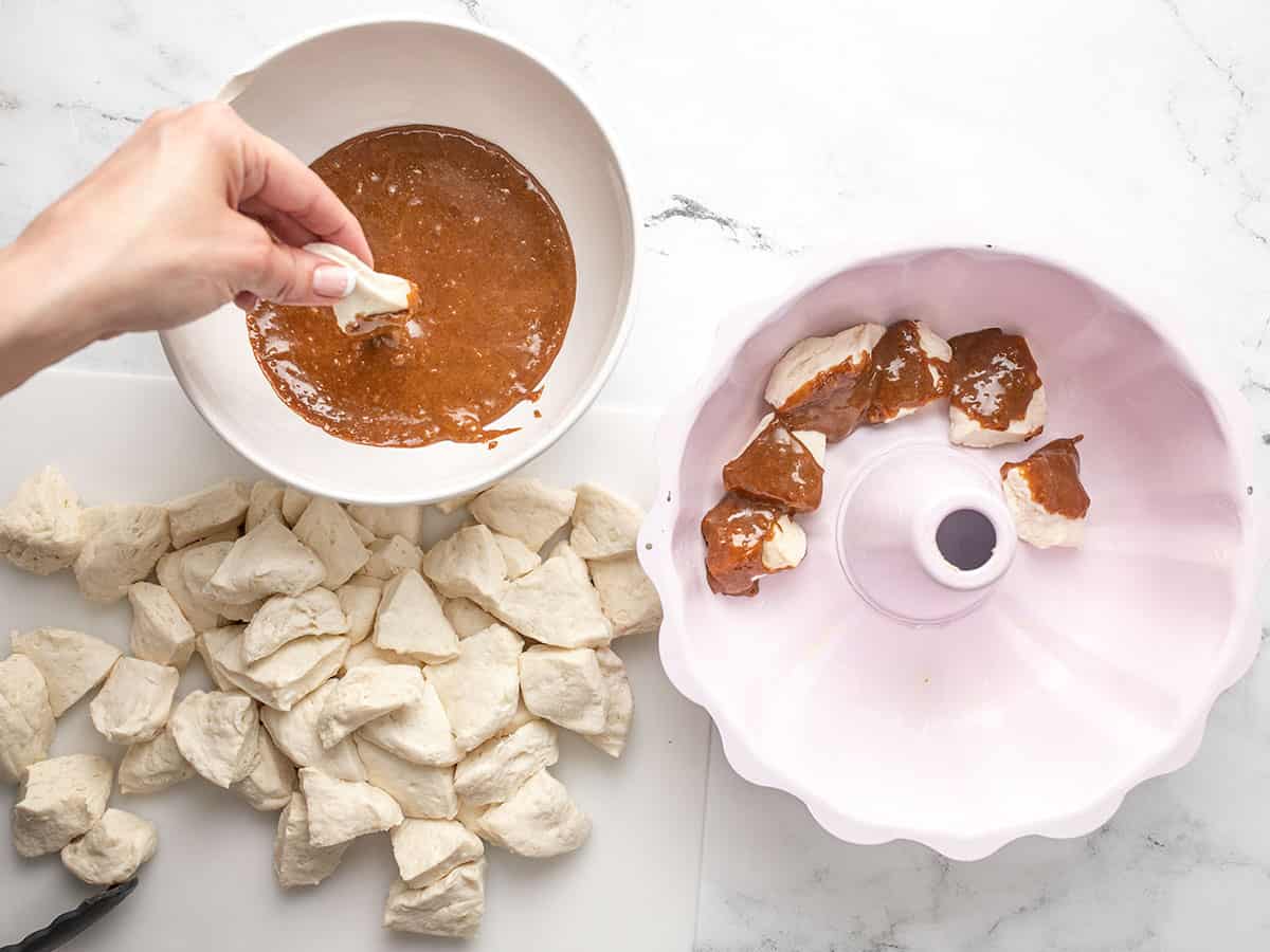 Overhead shot of hand dipping a biscuit piece into a bowl of caramel with pieces of biscuits and a Bundt pant next to it.