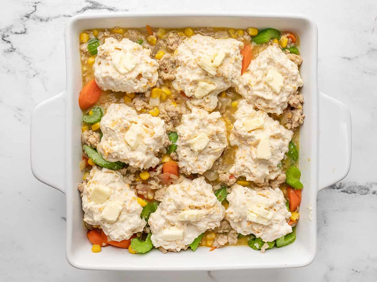 Overhead view of cooked chicken and biscuit casserole base in a white casserole dish with uncooked cheddar biscuits dropped on top. There are tiny pieces of butter on top of each biscuit. 