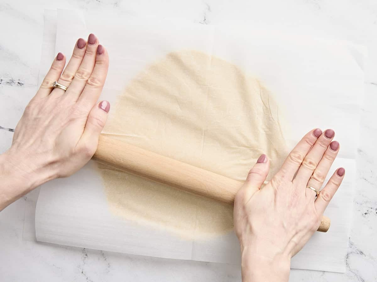 Hands rolling out sugar cookie dough between two sheets of parchment paper using a rolling pin.