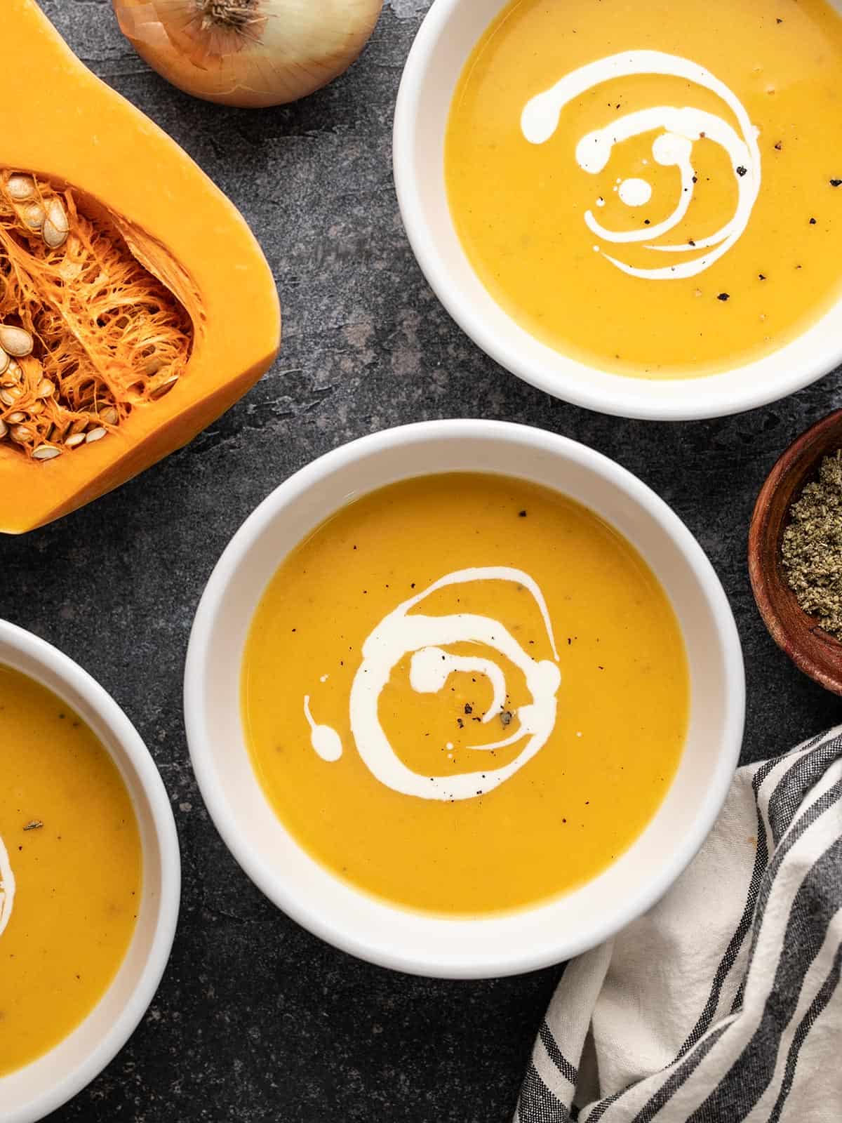 Three bowls of butternut squash soup next to a squash cut in half.