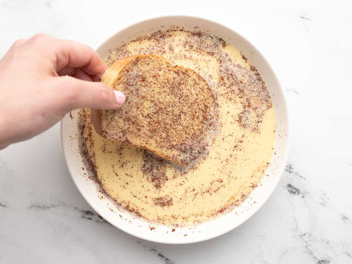 Hand dipping slice of bread into custard mixture in a white bowl.