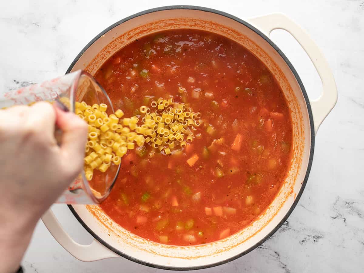 Hand pouring uncooked ditalini into a pot of simmering pasta e fagioli soup. 