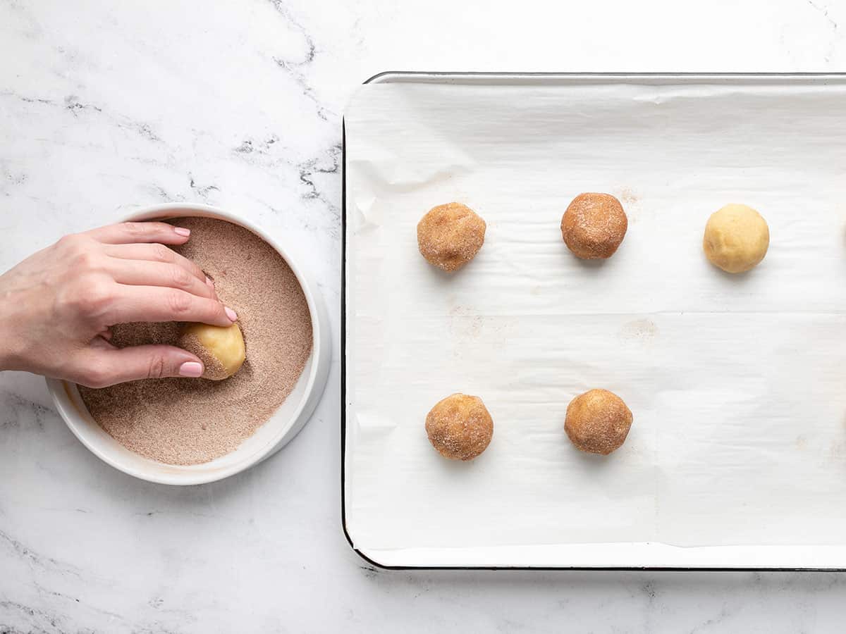 Balls of snickerdoodle dough being rolled in cinnamon sugar.