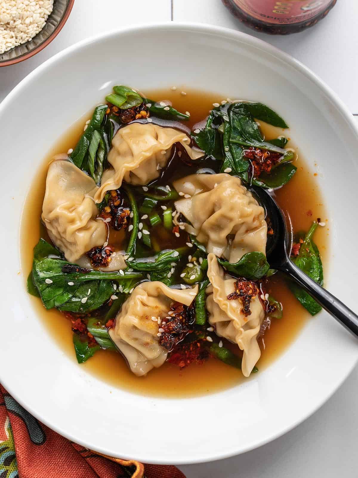 A close up view of dumpling soup in a white bowl topped with white sesame seeds and chili crisp.