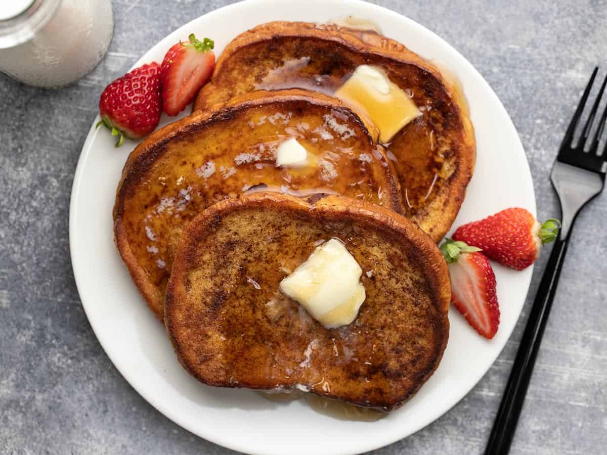 Overhead shot of three slices of cooked french toast on a white plate with sliced strawberries.