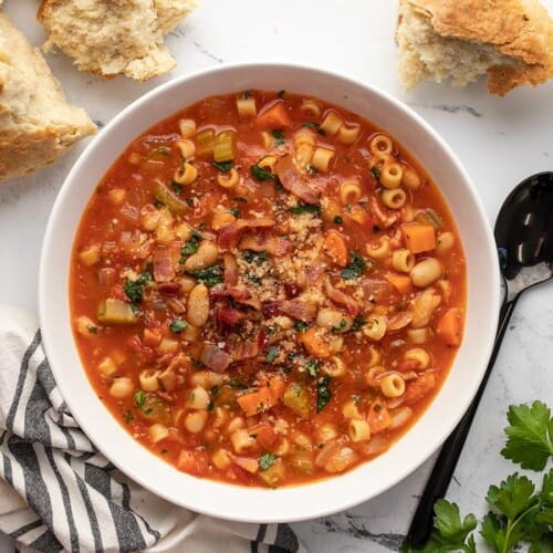 A white serving bowl of pasta e fagioli soup topped with bacon, parsley and parmesan cheese, and surrounded by torn bread, uncooked pasta, a black serving spoon, italian parsley and a decorative blue and white dish cloth.