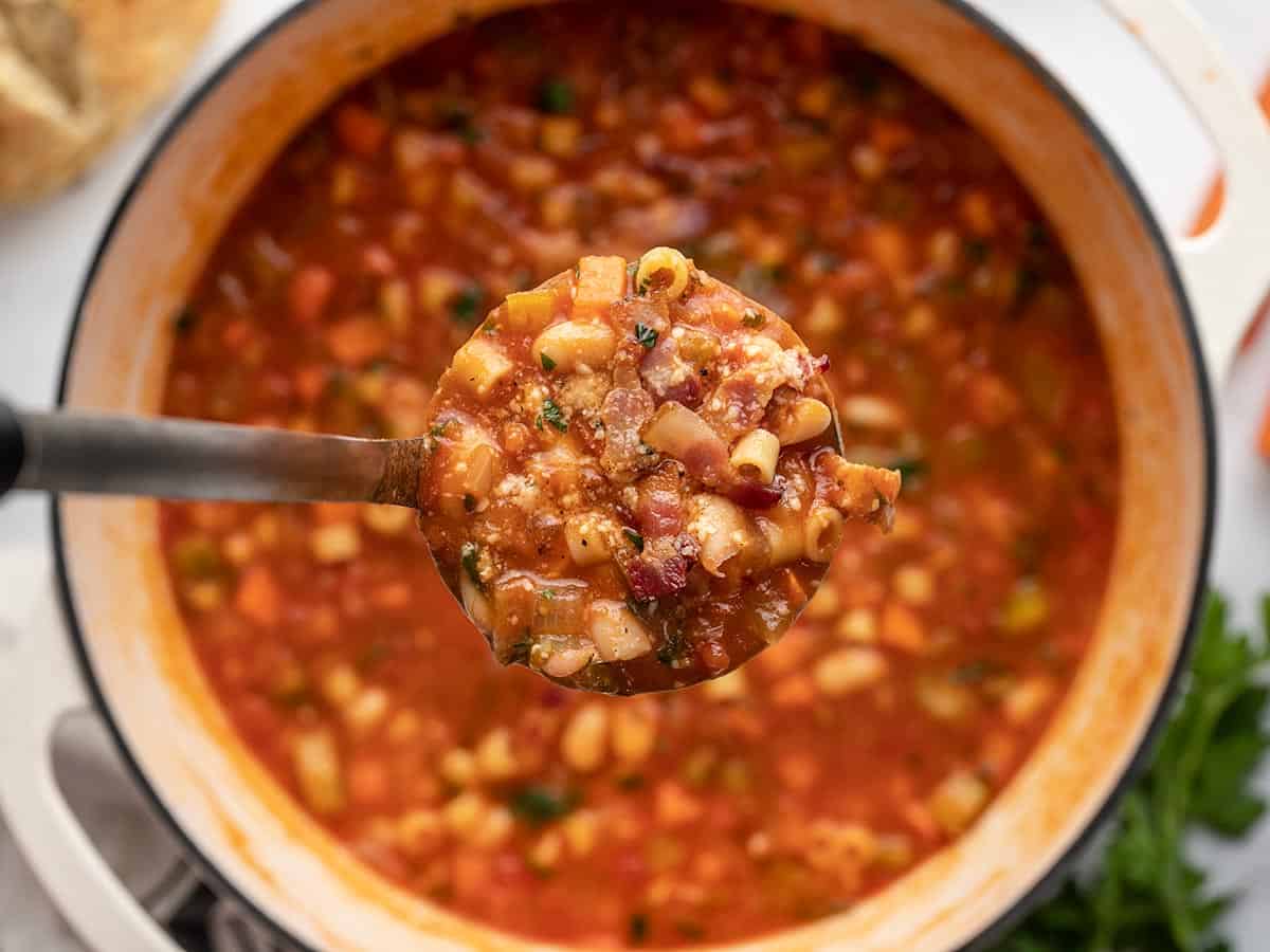 Close up of a ladle full of cooked pasta e fagioli soup and a large pot full of more soup in the background.