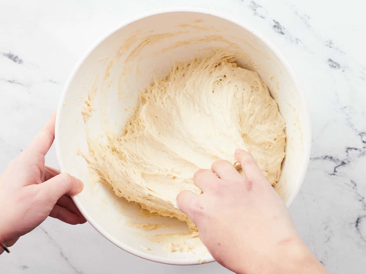 Ingredients for a no-knead focaccia style pizza crust after they have fermented for two hours in a white bowl. A left hand is stablizing the bowl with the right hand pulls the dough away from the side of the bowl. 