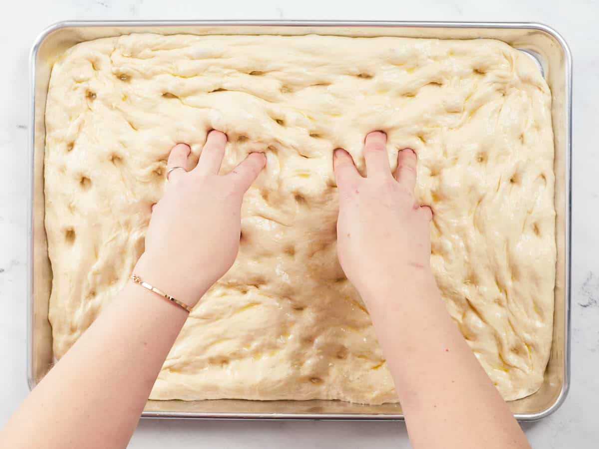 A raw pizza dough that has fermented for a second time on a rimmed baking tray, two hands are pressing dimples into the center of the dough. 