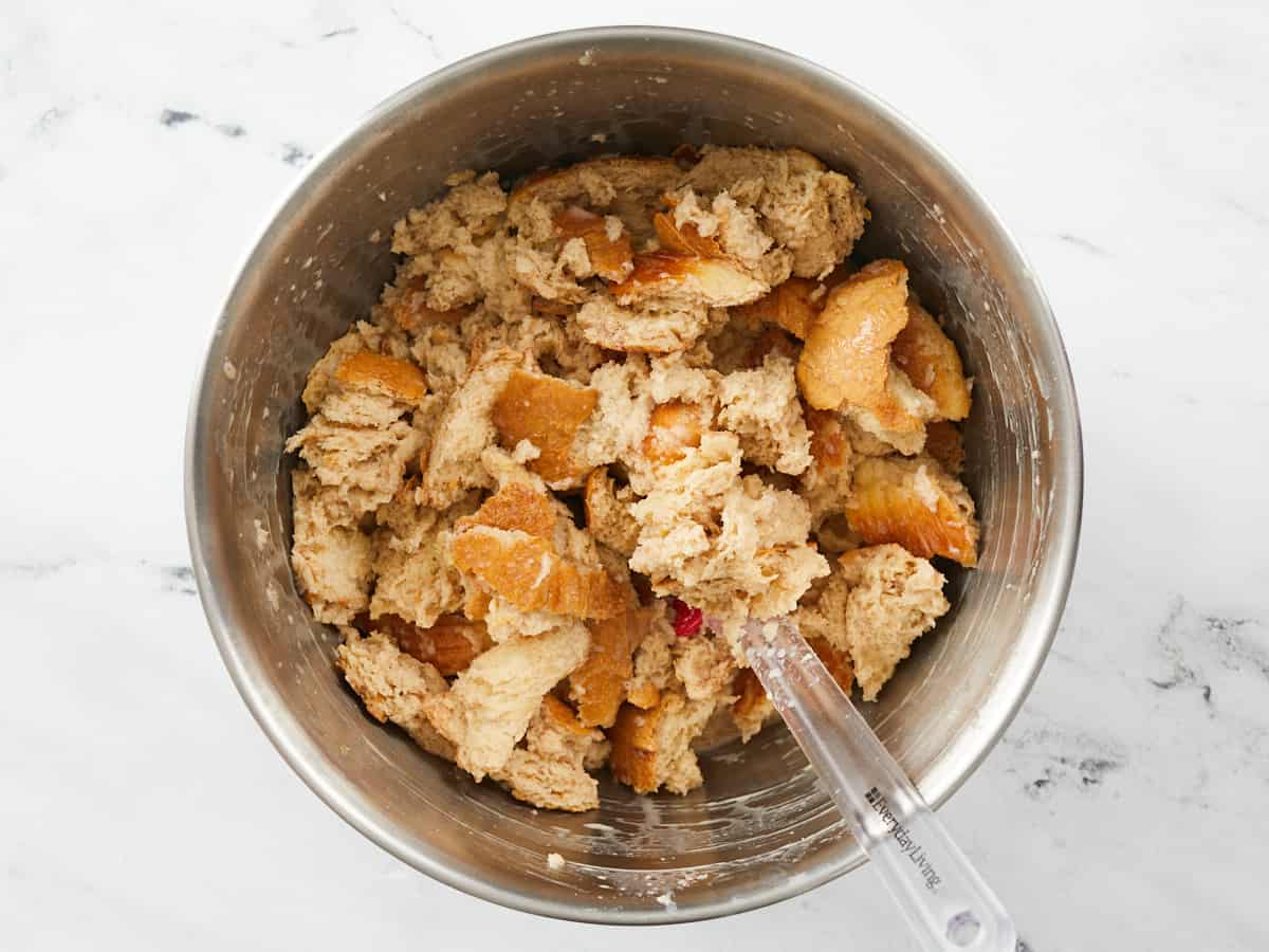 A large metal mixing bowl filled with torn pieces of bread that have been tossed in a custard mixture and left to soak with a red spatula sitting in the bottom right corner of the bowl. 