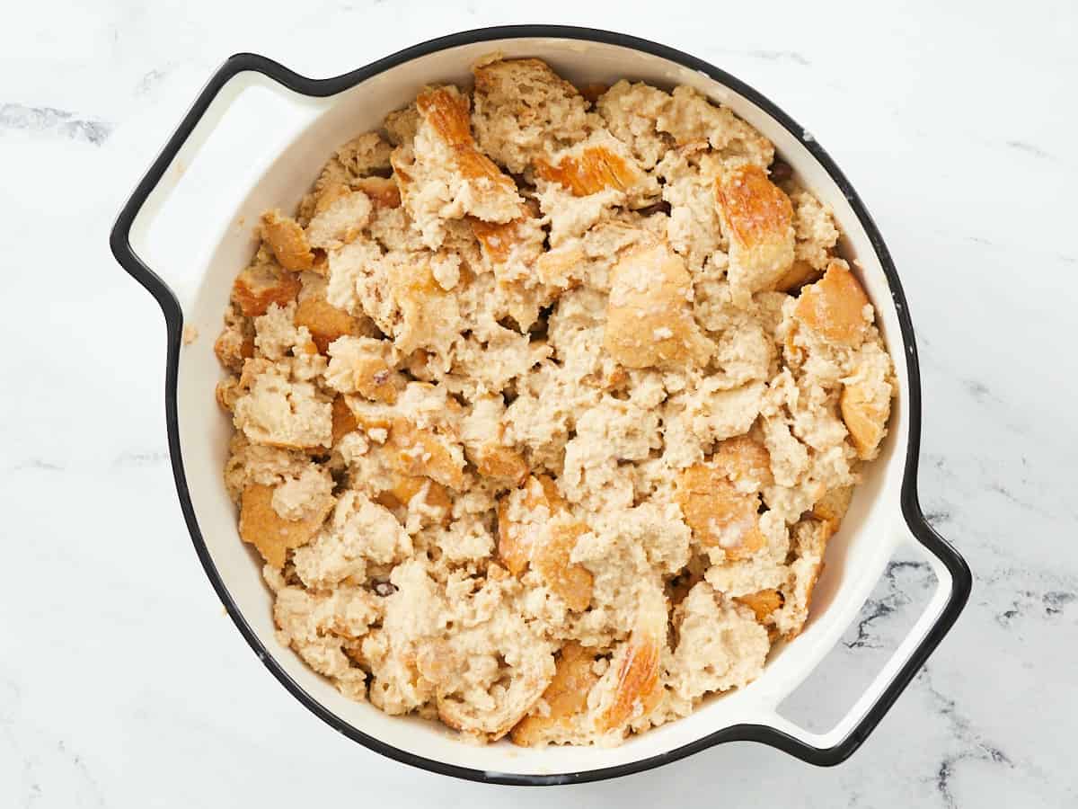 A round, white enamel baking dish with a black rim on a white marble background that is filled with an unbaked bread pudding. 