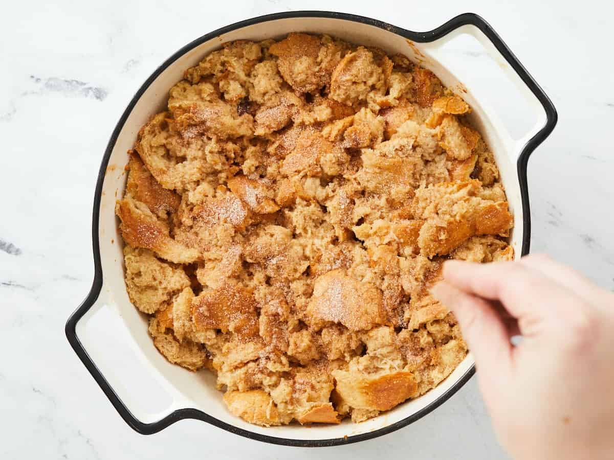 A round white enamel baking dish on a white marble background that is half baked. A hand in the lower right corner of the frame is sprinkling cinnamon sugar on top of the casserole.