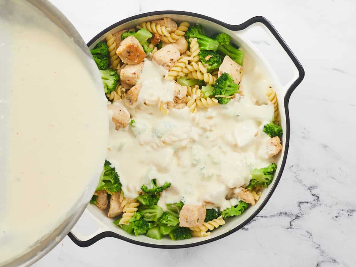 A circular, white enamel cast iron with a black rim sitting on a marble background. The casserole dish is filled with a mixture of cooked pasta, cooked chunks of seasoned chicken breast, and broccoli florets. Homemade alfredo sauce is being poured over the mixture from a stainless steel skillet raised above the casserole from the left side of the frame. 