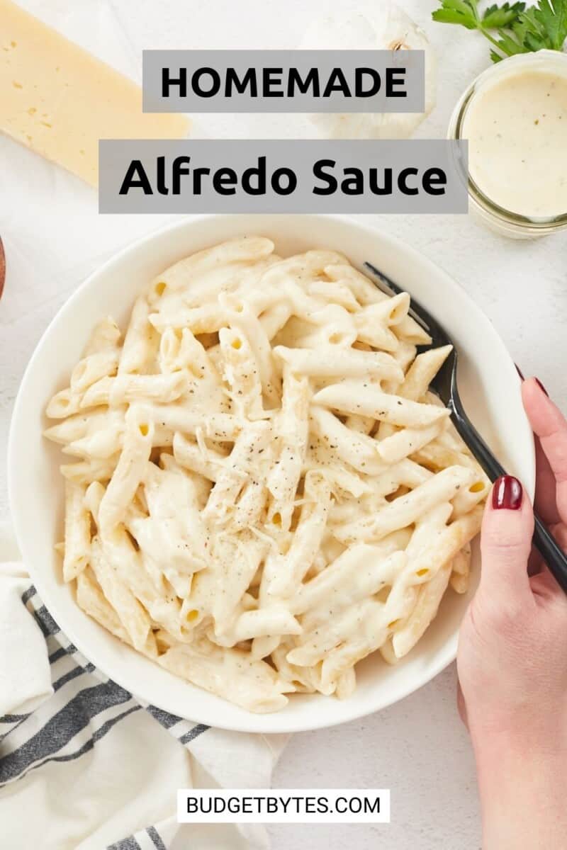 A large, vertical image of a bowl of penne pasta tossed in creamy homemade alfredo sauce. A hand is holding the right side of the bowl and a black spoon that's resting in the dish on the right side. The bowl is surrounded, from left to right, with a white and gray striped dish cloth, a block of parmesan cheese, some sprigs of parsley and a mason jar filled with alfredo sauce. Over the top center of the image is a gray text box with black text that says "Homemade Alfredo Sauce."