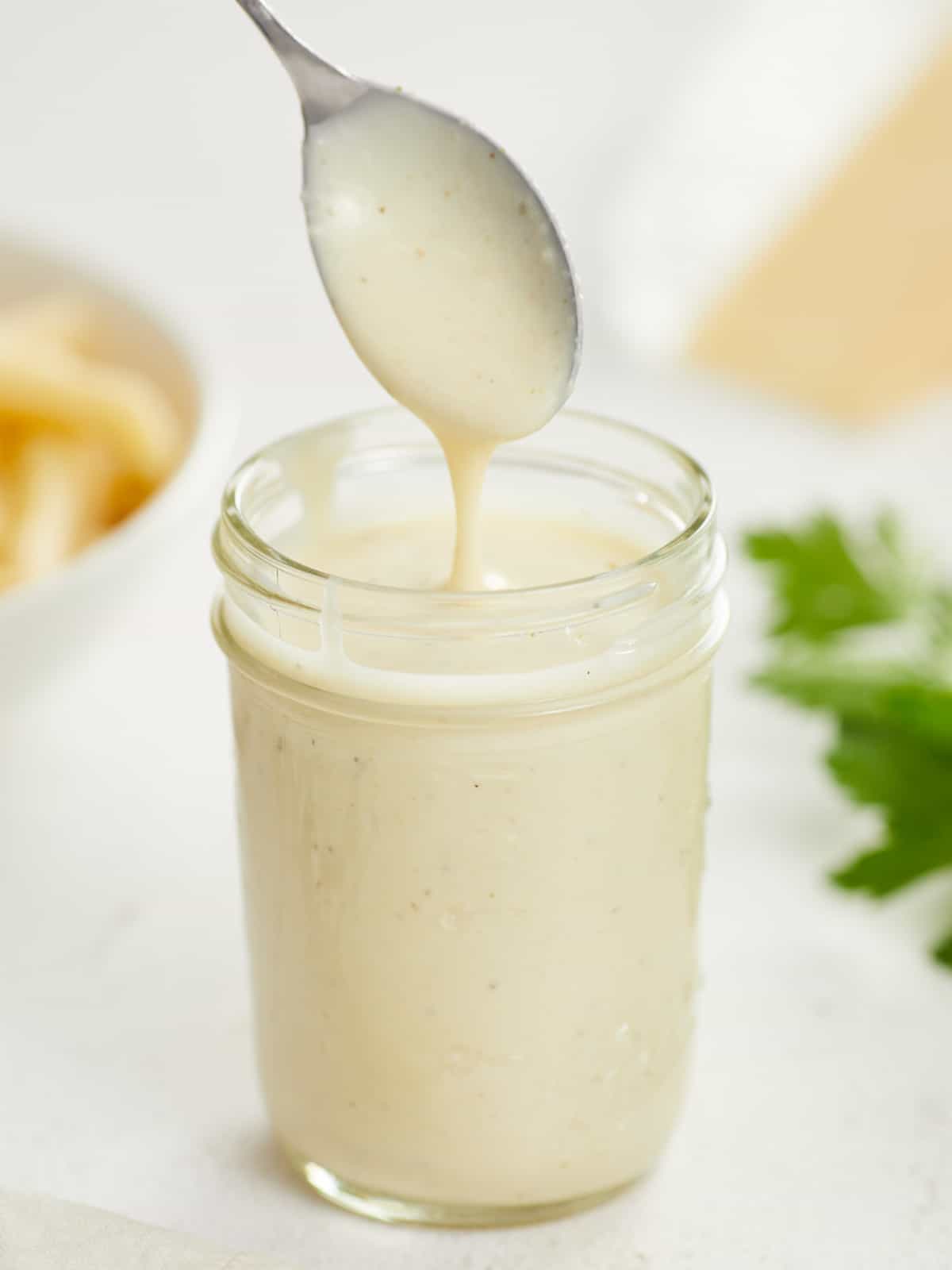 A mason jar filled with alfredo sauce and a metal spoon raised above the jar after it's been dipped into the sauce so it's dripping off, and blurred in the background is a block of parmesean cheese, a bowl of uncooked pasta and some fresh italian parsley.