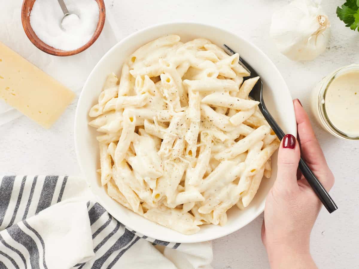 A large white bowl filled with cooked penne pasta dressed with homemade alfredo sauce, a hand is holding the right side of the bowl and a black metal fork, various props surround the bowl including a grey striped dish cloth, a block of parmesan cheese, a wooden bowl of salt and a mason jar of alfredo sauce.