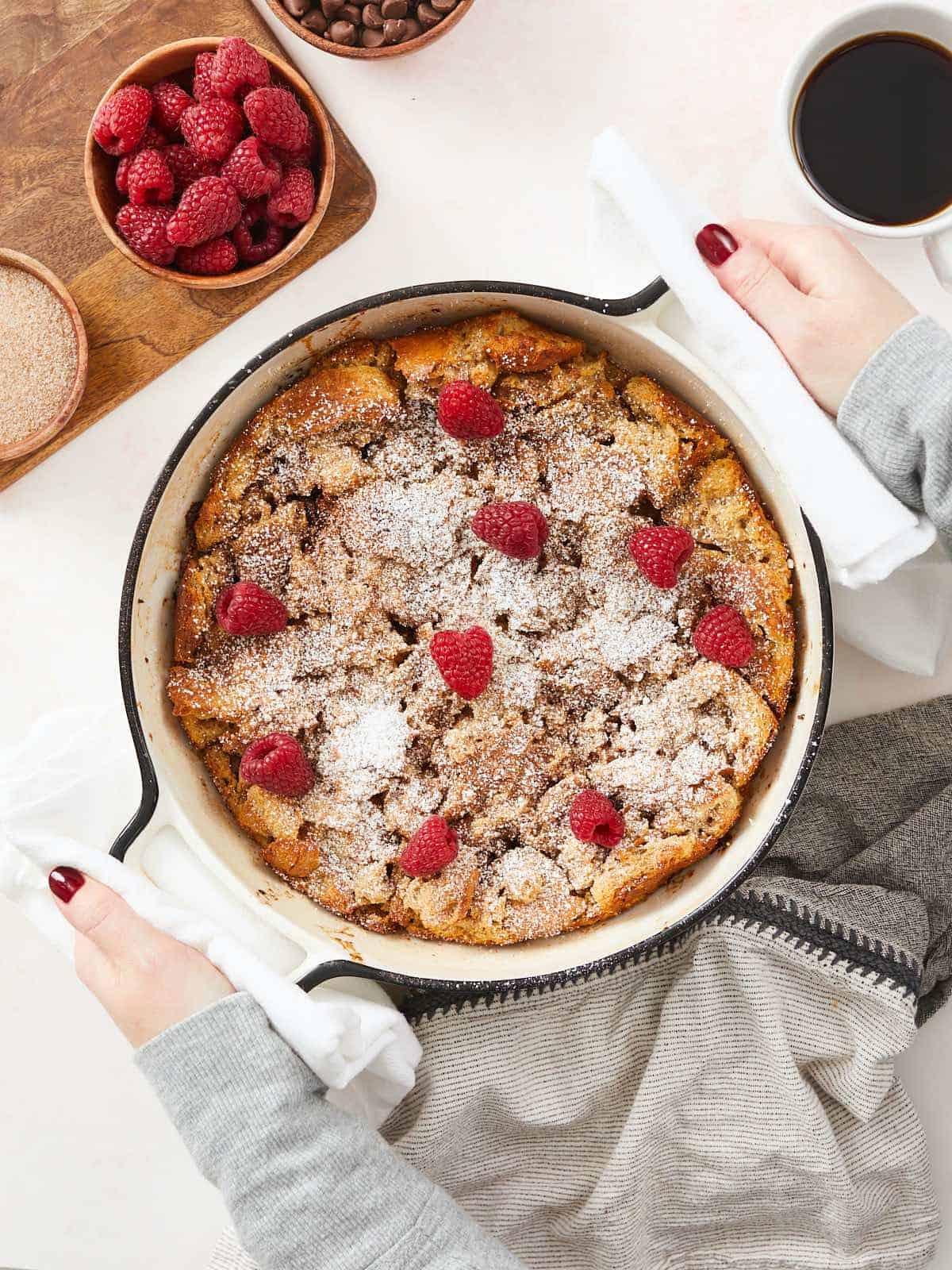 A vertical image of a baked bread pudding in round, white enamel cast iron baking dish topped with powdered sugar and fresh raspberries. There is a cutting board in the top left corner of the frame with a wooden bowl of raspberries and next to it is a cup of coffee partially out of frame.