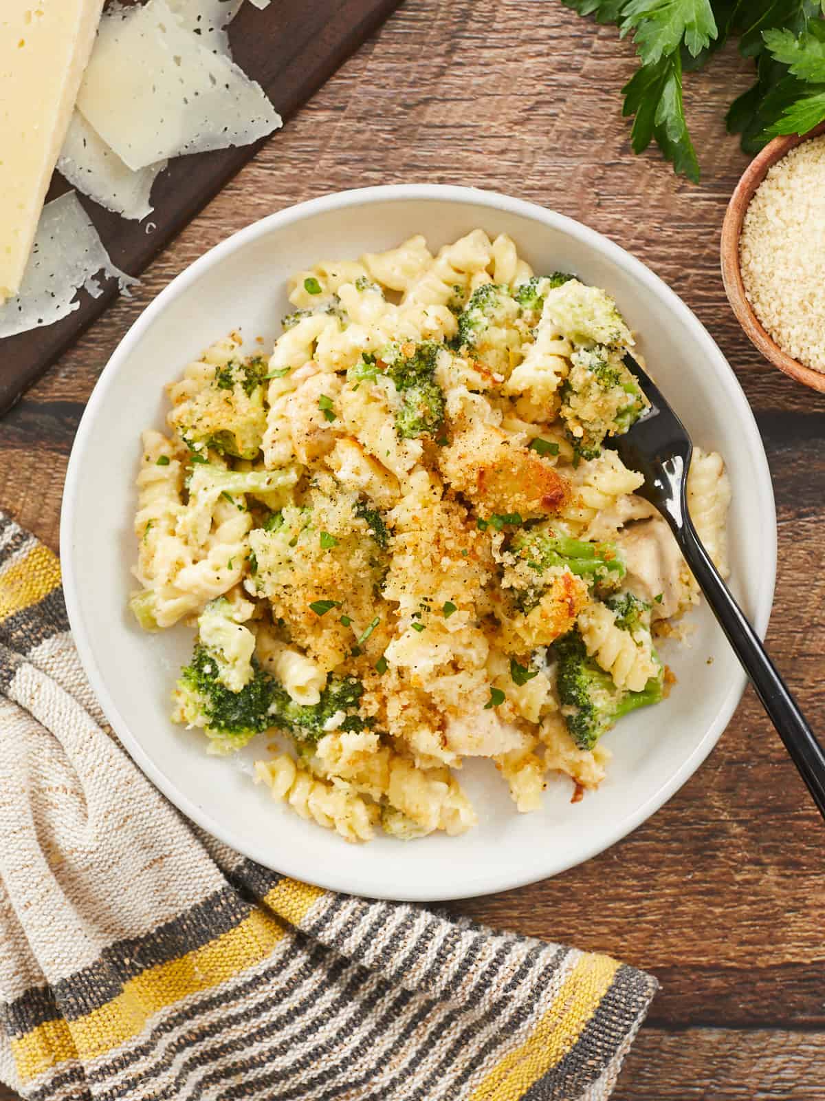 A large serving of chicken alfredo bake in a flat, white bowl, and a black fork is resting on the right side of the bowl. The bowl is sitting on a wood background surround with a yellow and gray striped dish cloth, a dark wooden cutting board with Parmesan Cheese shavings on it, and a sprig of parsley and a bowl of untoasted breadcrumbs.