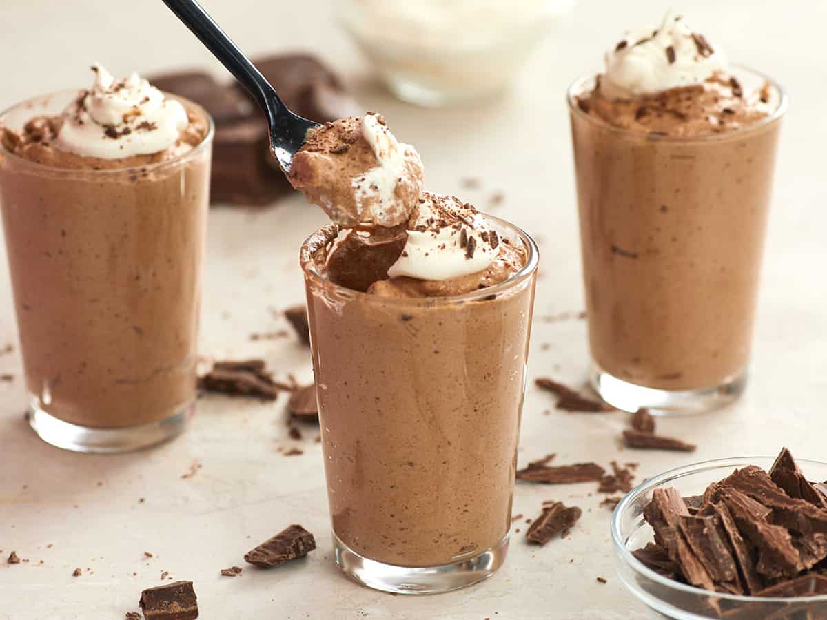 A spoon digging into a cup of chocolate mousse with whipped cream. 