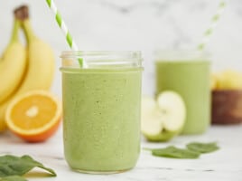 Side shot of green smoothies surrounded by cut fruit.