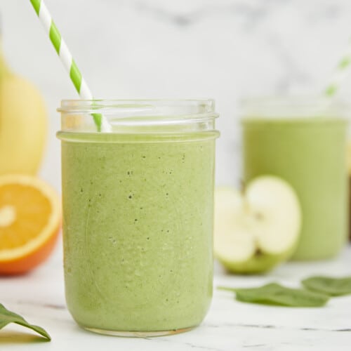 Side shot of green smoothies surrounded by cut fruit.