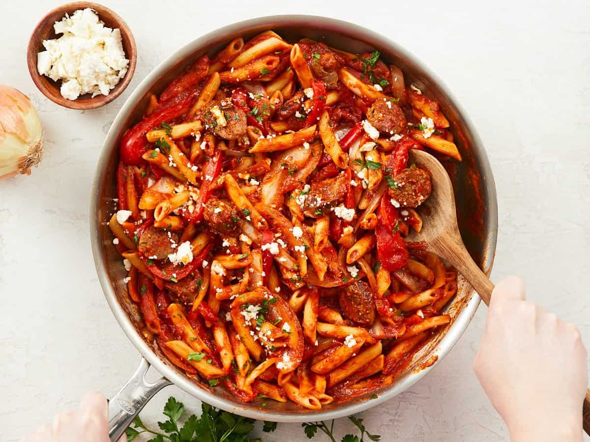 Overhead view of a skillet with sausage and peppers being stirred with a wooden spoon.